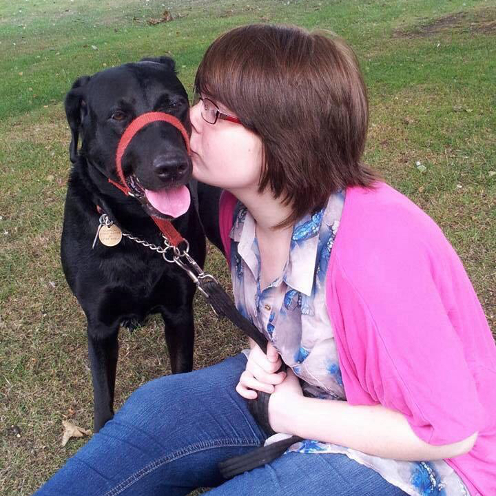 Ami is sitting on the grass, with Millie standing next to her. Millie is a black Doberman mixed Labrador. She is happy as Ami is giving her a kiss on her cheek