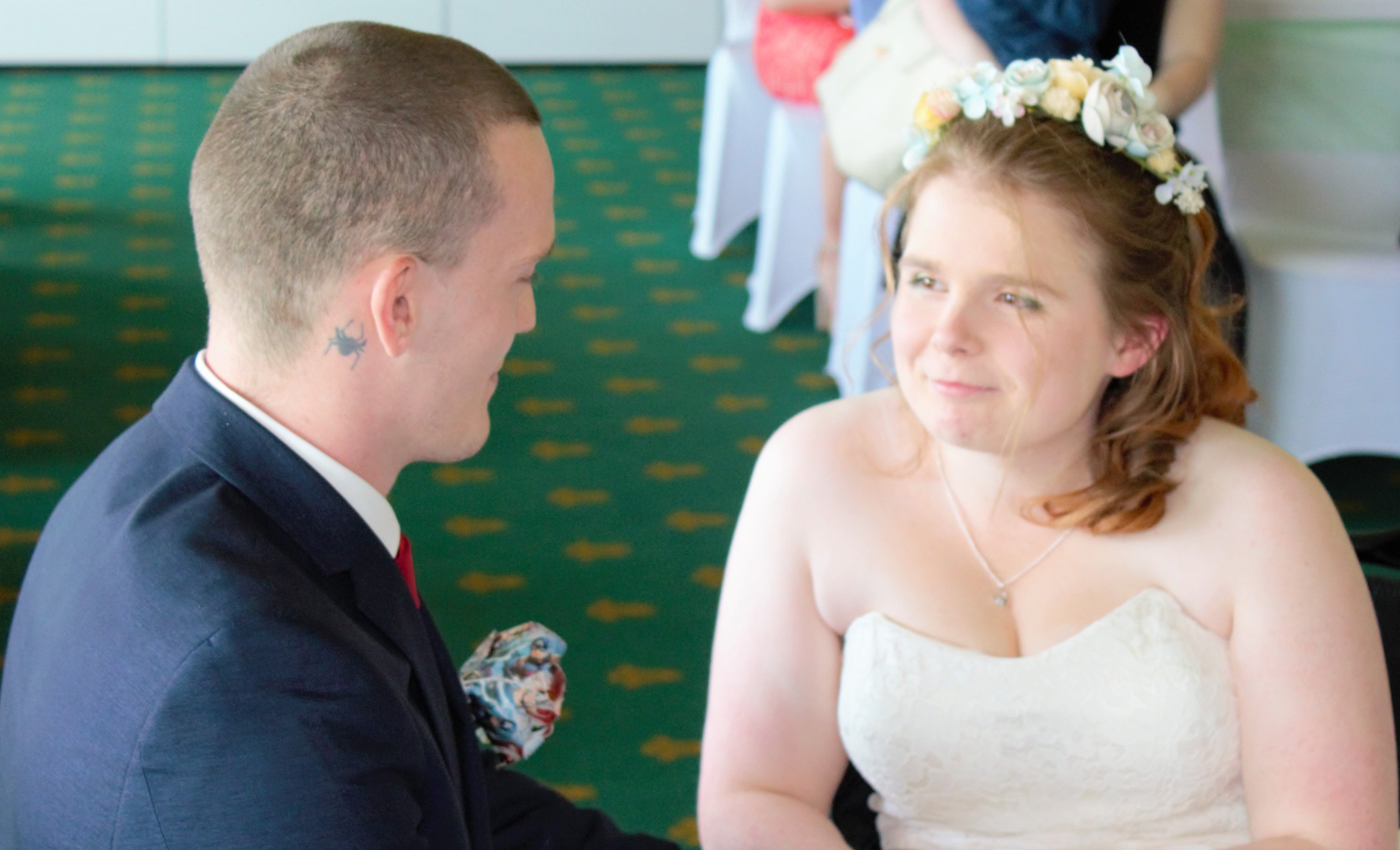 Ami and Ewan are declaring their vows. Ami is sitting in her wheelchair, wearing an ivory satin knee-length dress with a lace bodice. Ewan is kneeling beside her. He is wearing a navy suit and a white shirt, he also has a comic book page styled rose buttonhole.