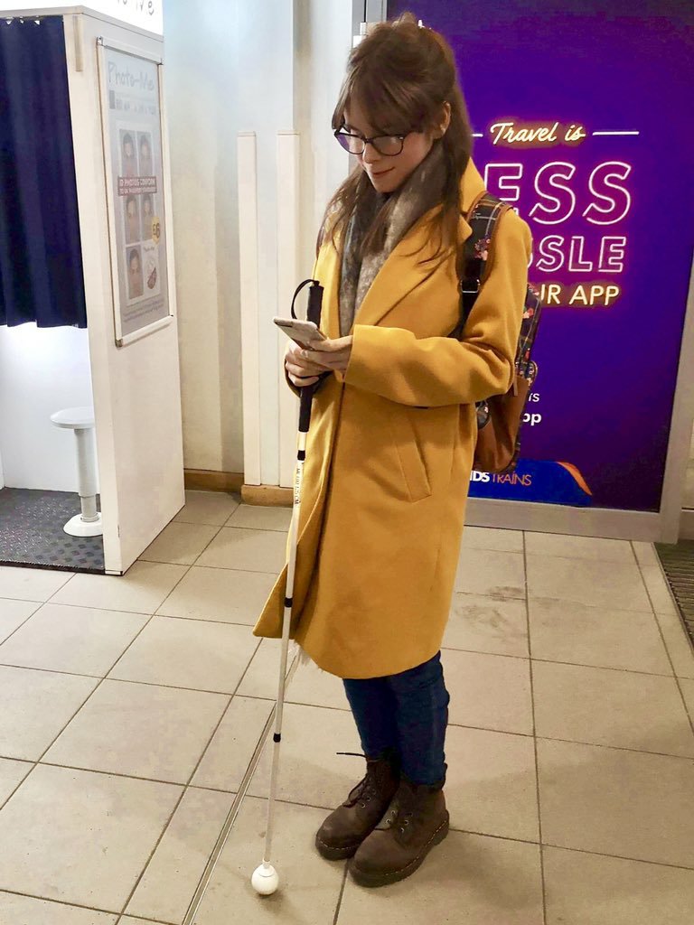 Chloe is wearing a knee-length yellow coat with beige grey scarf and backpack over her left shoulder. Also dark blue denim jeans brown boots. Chloe standing, holding long white cane whilst using phone.