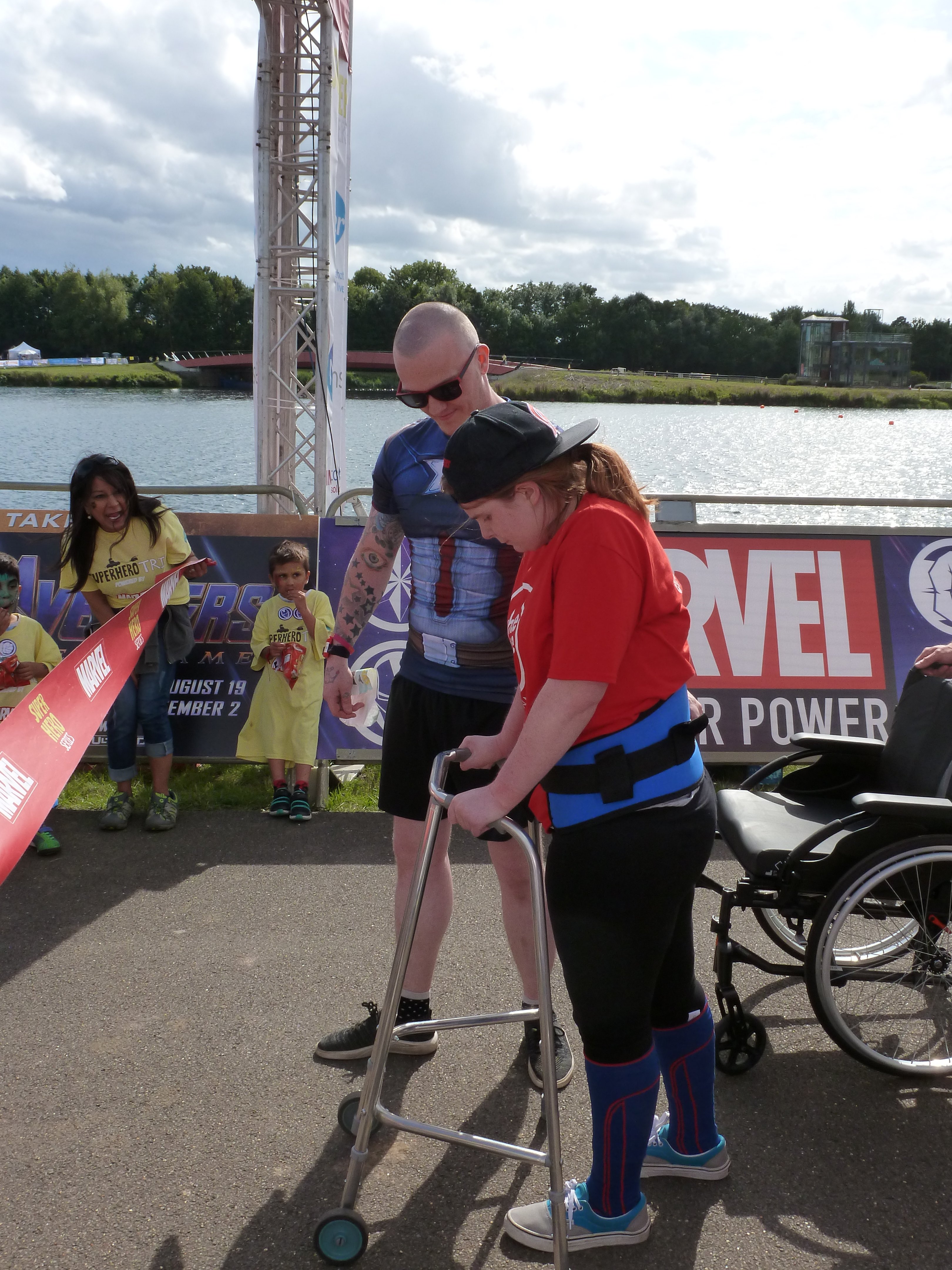 Ewan is wearing a Captain America torso shirt with black shorts, trainers and sunglasses. I am red t-shirt The White Lodge logo on the front, leggings, blue lycra garments which aid my walking ability, grey Vans. Also, cap back to front. have transfer belt around waist allows Ewan guide me, should lose balance. taking step forward frame about cross finish line assisting me. This photo was taken at Superhero Tri, Dorney Lake in Windsor.