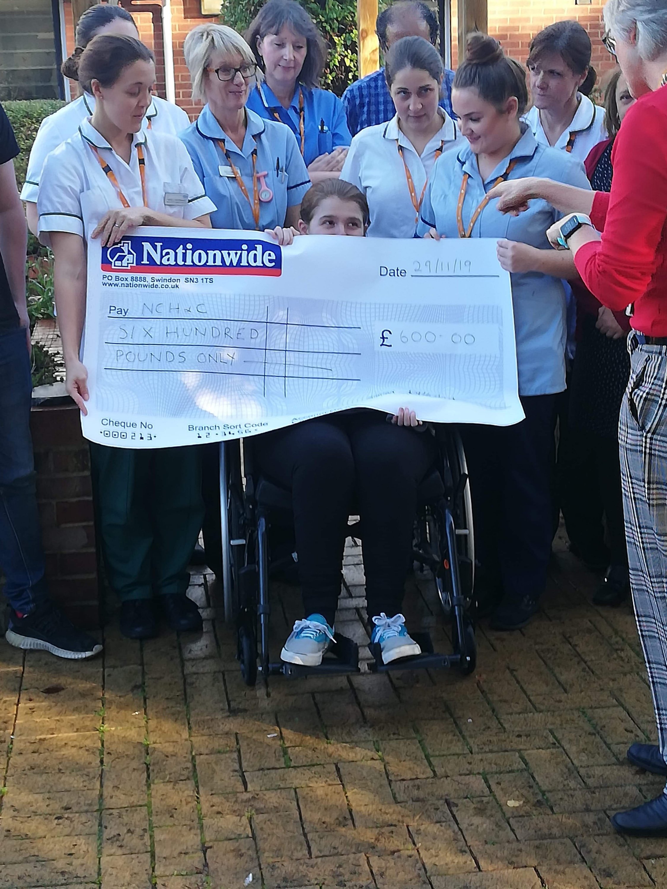 Ami and Ewan presenting a cheque which was made from the donations of their wedding guests. Ami is trying to hide behind the cheque as her rehab doctor is praising her.