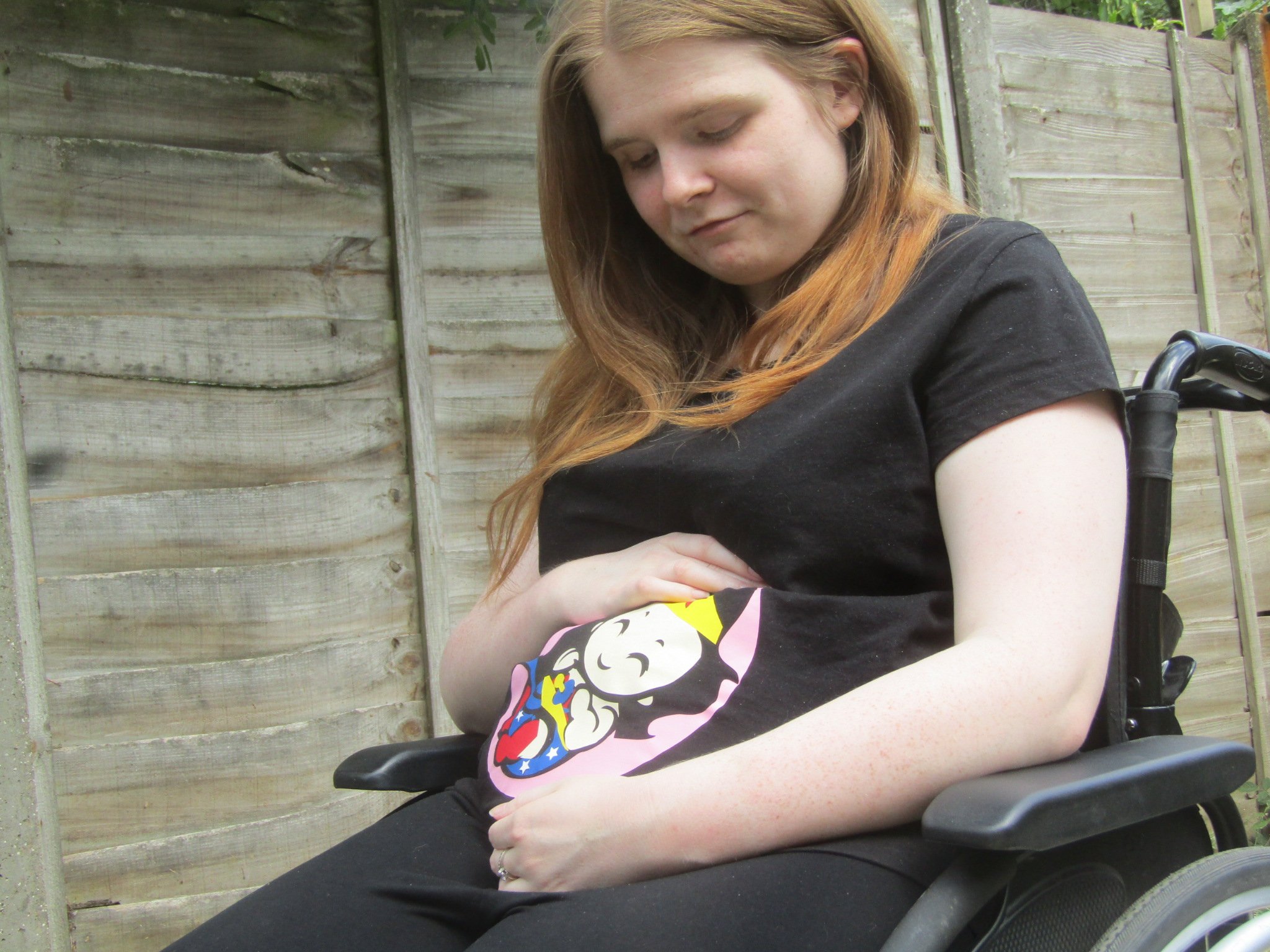 Close up side view version - Ami is sitting in her wheelchair in the garden. She is wearing a black t-shirt with a Wonder Woman baby design which covers the bump area. Ami is looking down and smiling while cradling bump.
