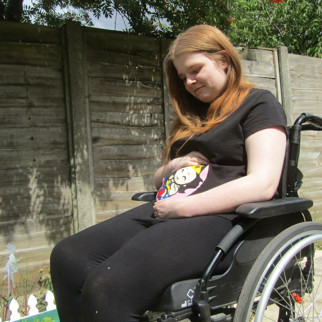 Side view version - Ami is sitting in her wheelchair in the garden. She is wearing a black t-shirt with a Wonder Woman baby design which covers the bump area. Ami is looking down and smiling while cradling bump.