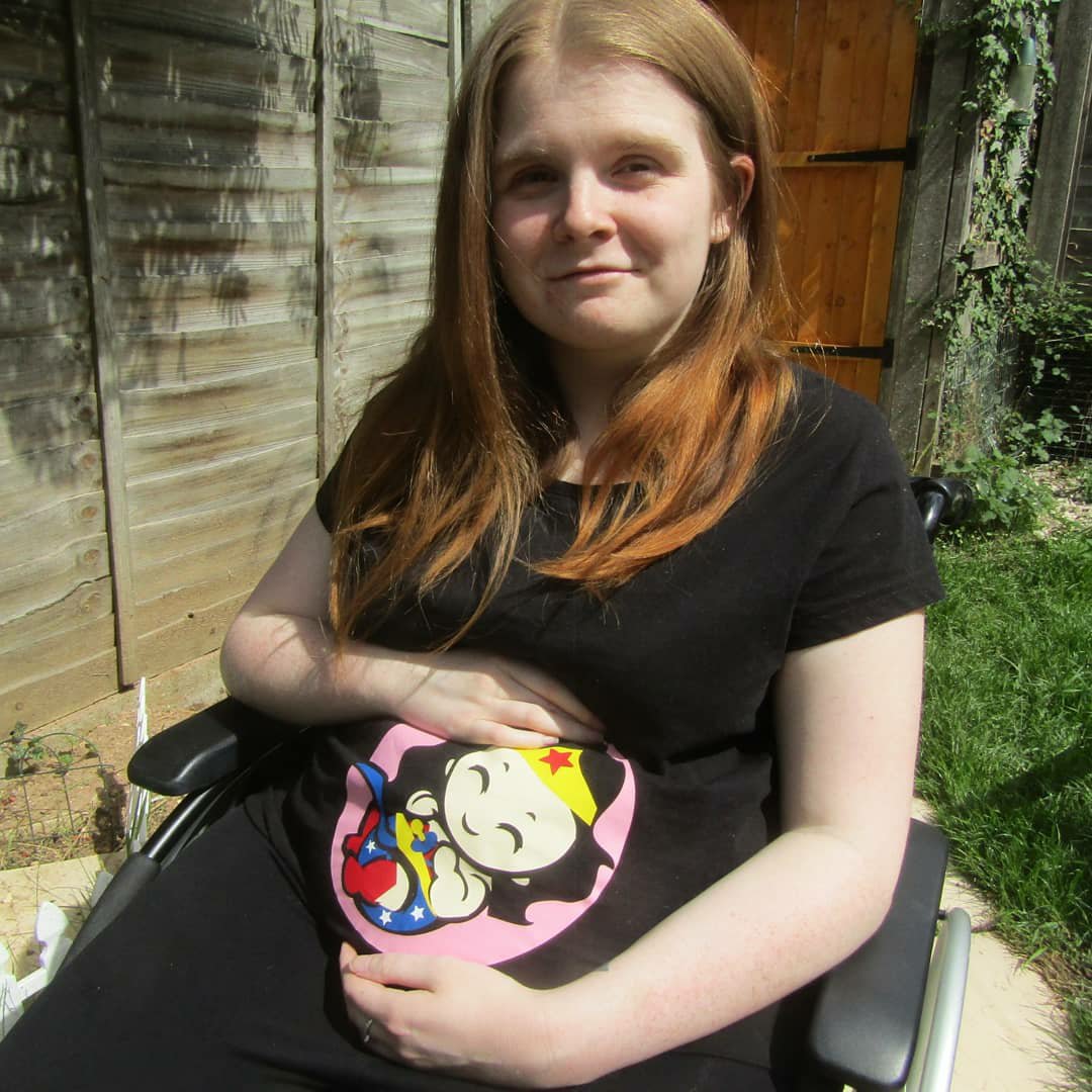 Close up version- Ami is sitting in her wheelchair in the garden. She is wearing a black t-shirt with a Wonder Woman baby design which covers the bump area. Ami is looking at the camera and smiling while cradling bump.
