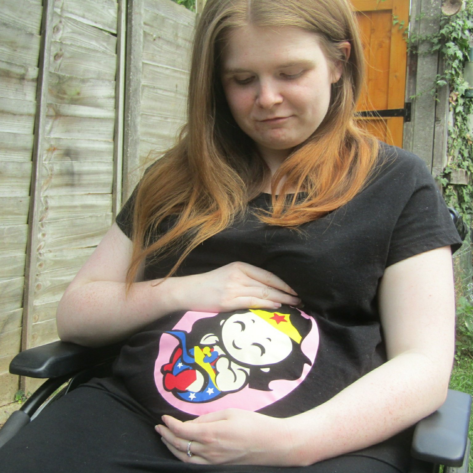 Close Up Version- Ami is sitting in her wheelchair in the garden. She is wearing a black t-shirt with a Wonder Woman baby design which covers the bump area. Ami is looking down and smiling while cradling bump.
