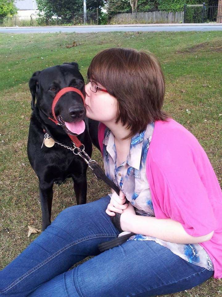 A photo of younger Ami with her dog Millie. Millie is a black Doberman cross Labrador. Ami is sitting on the grass giving Millie a big kiss on her cheek.