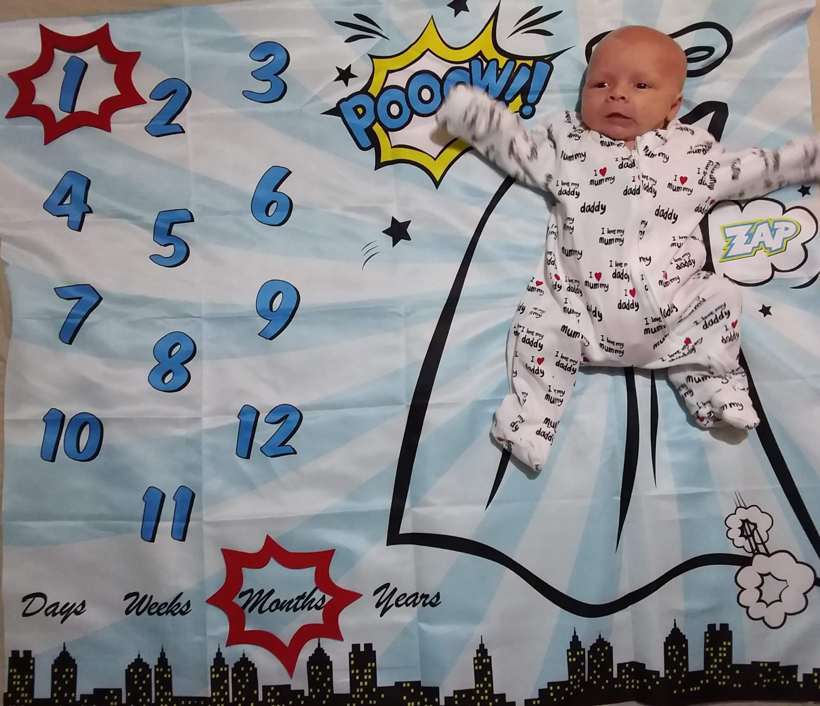 Photo of Daisy on her superhero themed milestone blanket with caption shapes around the number 1, and the word months.