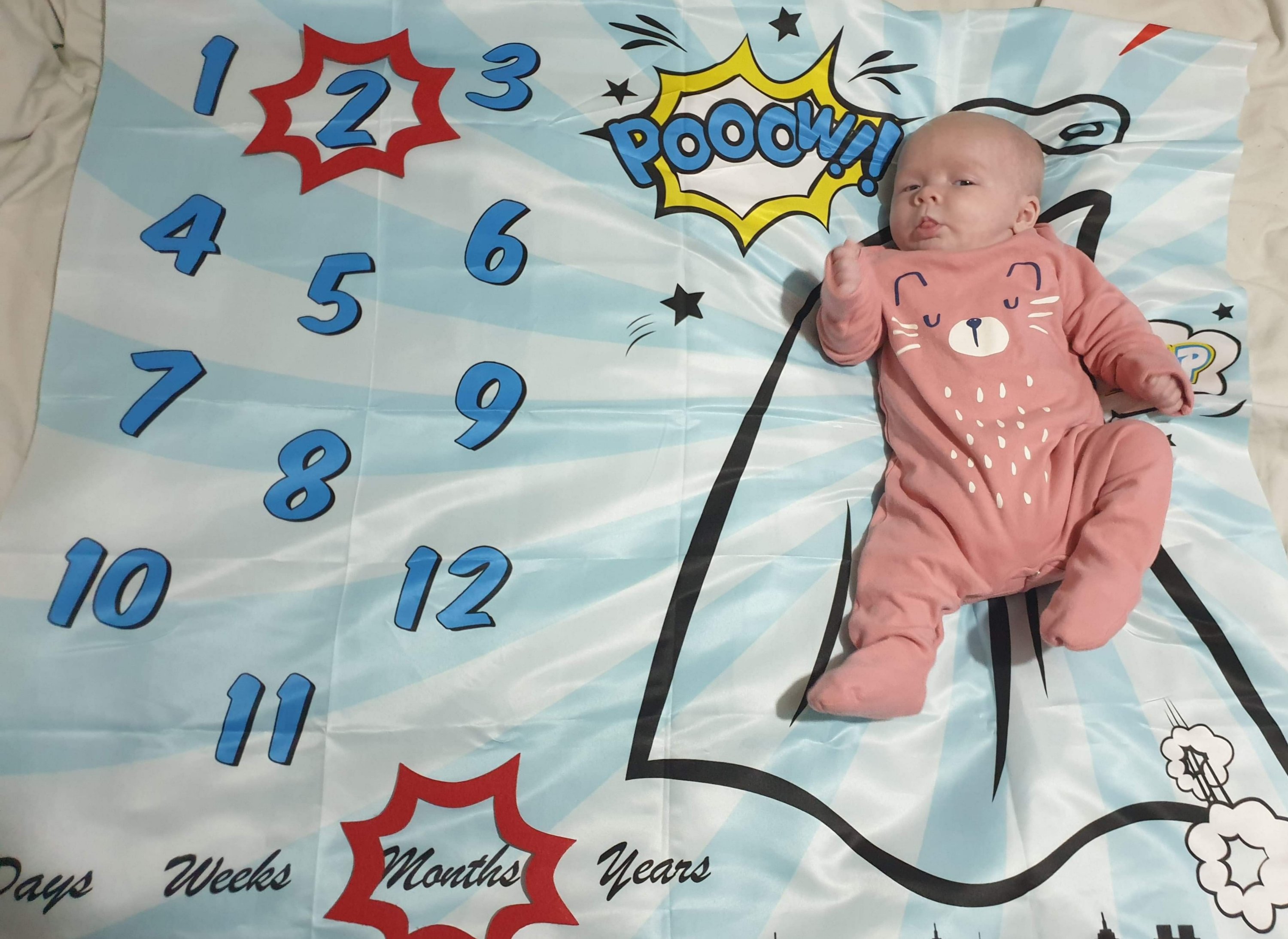 Photo of Daisy on her superhero themed milestone blanket, with a red caption shape around the number 2 and another shape around the word months