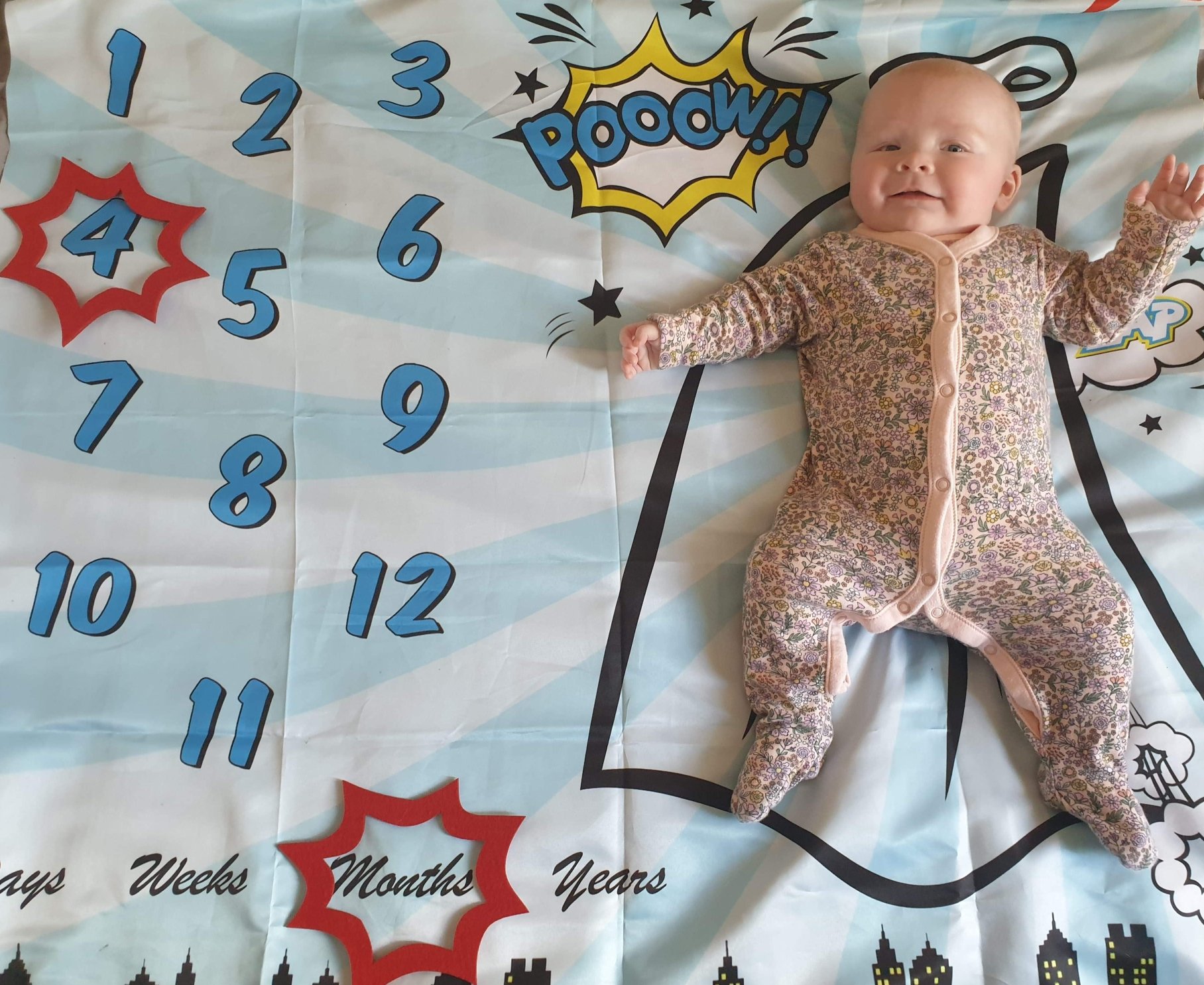 Photo of Daisy on her superhero styled milestone blanket, with one red caption shape around the number 4, and the other red caption shape around the word months.