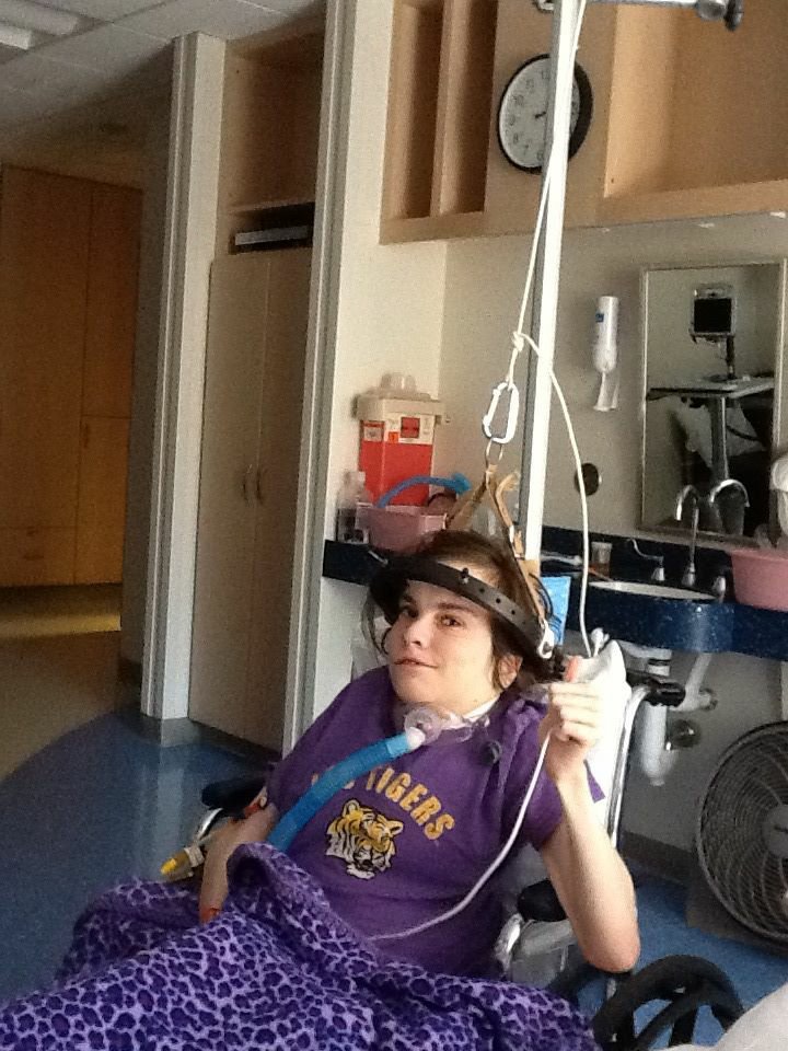 Post-surgery photo of Jamie, she is sitting in her chair, whilst wearing the Halo Traction device on her head, has a breathing tube attached to her tracheostomy, and has intravenous pain medication and food attached to a portable pole beside her.