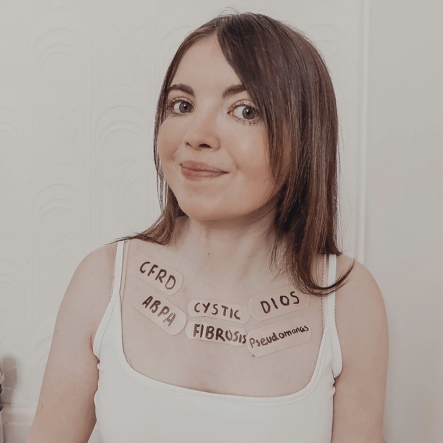 Lucy is looking at the camera, smiling. She has 6 clear stickers attached to her chest area, one sticker for each condition she has. The sticks read, Cystic Fibrosis, CFRD (Cystic Fibrosis Related Diabetes), ABPA (Allergic Bronchopulmonary Aspergillosis), DIOS (Distal Intestinal Obstruction Syndrome, and Pseudomonas.