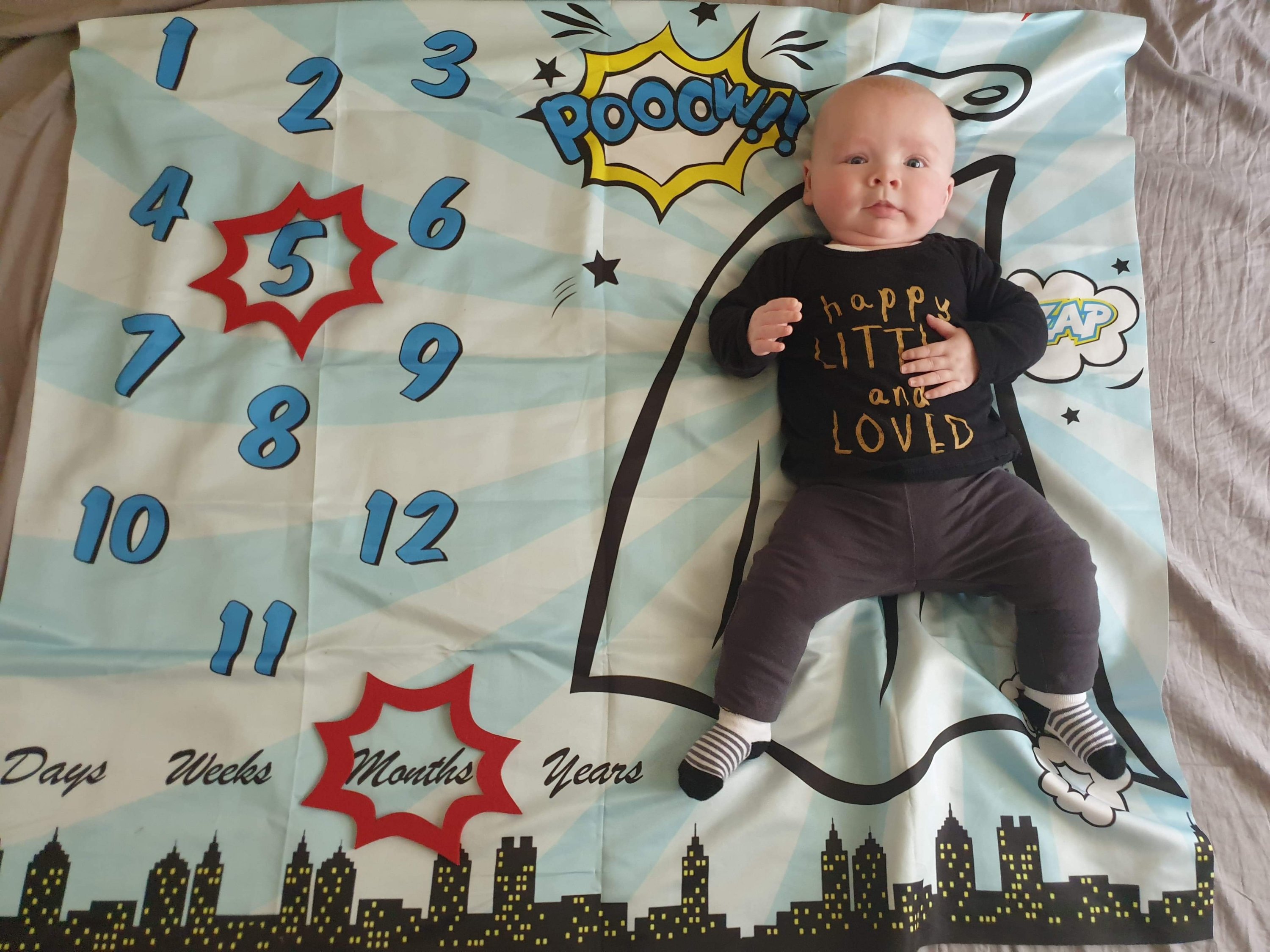 Daisy is laying on her superhero themed milestone blanket, with a red caption shape around the number 5, and another around the word months