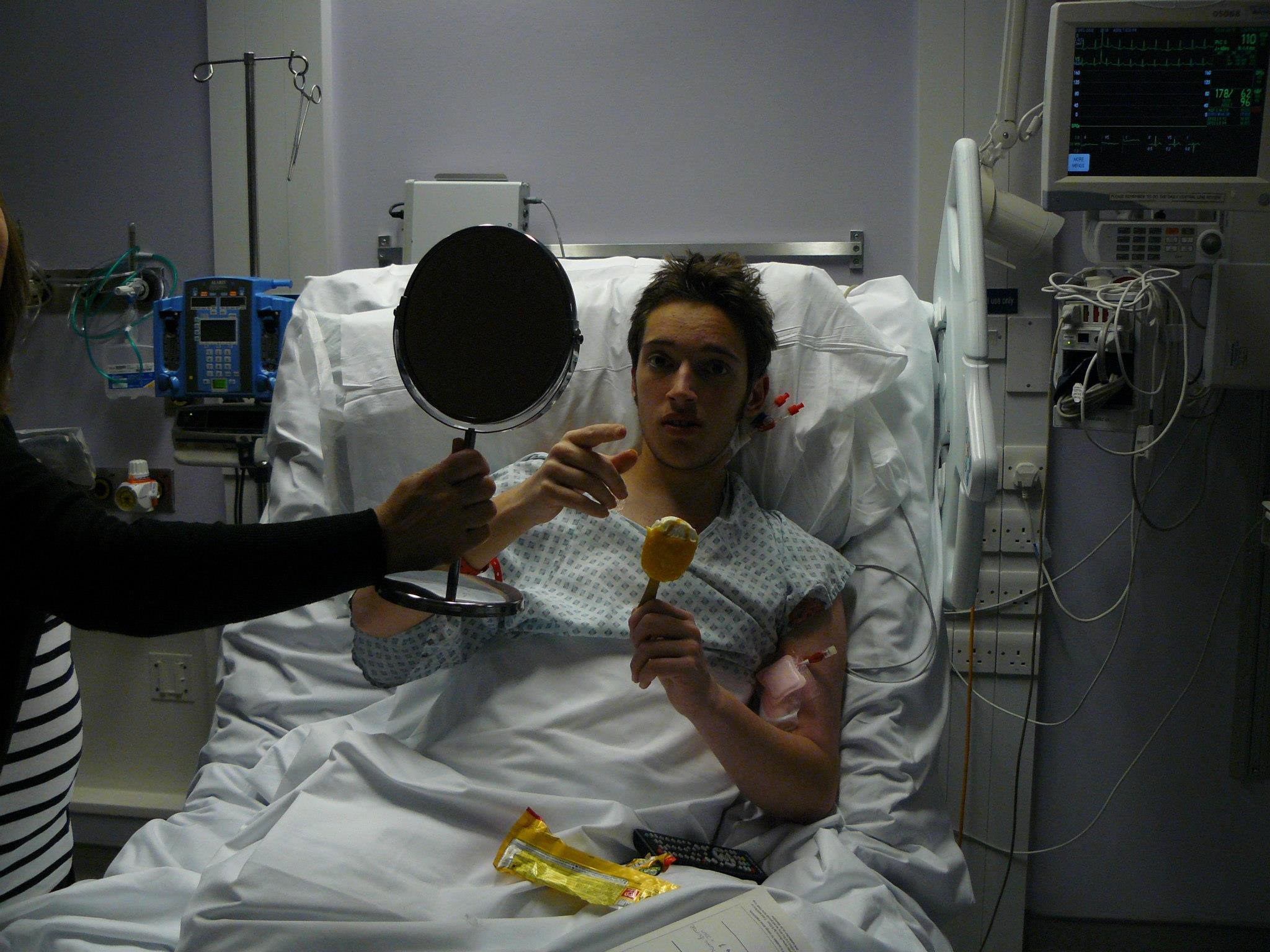 David is sitting up in a hospital bed, holding a brush and another person is holding a mirror in front of him. David has several tubes attached.