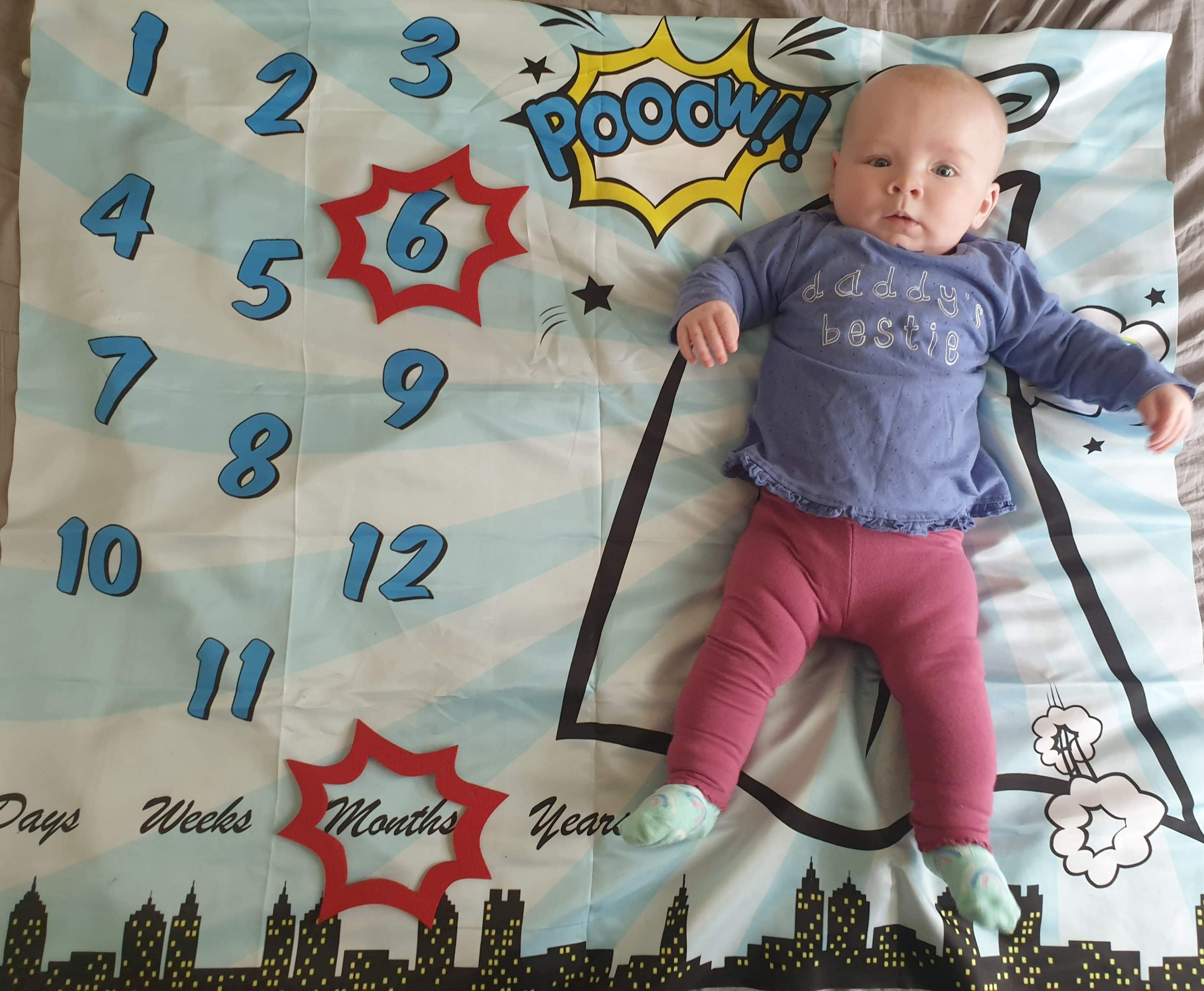 Daisy is laying on her superhero themed milestone blanket, with a red caption shape around the number 6 and the word months