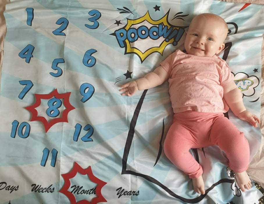 Daisy is smiling, laying on her superhero themed milestone blanket. There is a red caption shape around the number 8 and another caption shape around the word months
