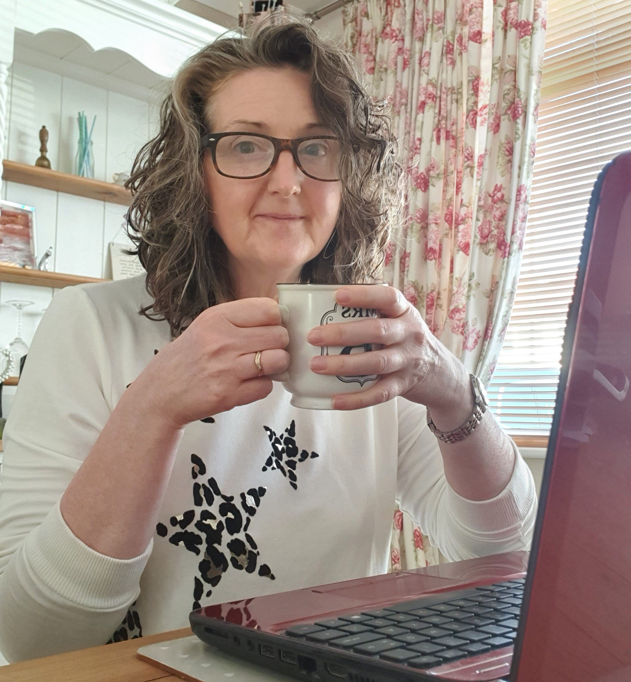 Tracy is sat at a table, in front of her laptop, holding a cup. Tracy has shoulder-length curled brown hair, is wearing glasses, and a white jumper with leopard-printed stars. She is gently smiling to the camera.