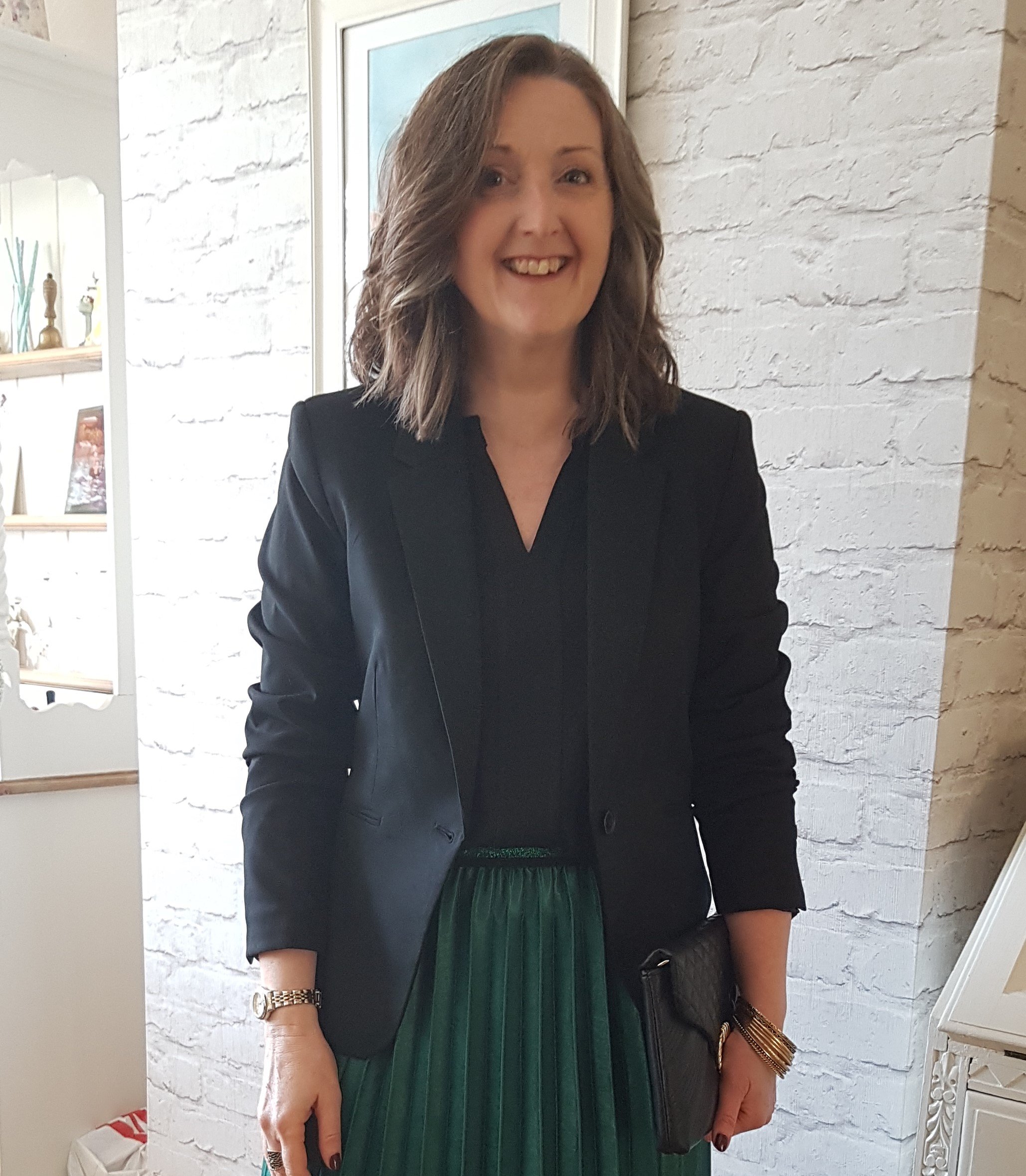 Tracy is standing at an angle in front of a white rustic wall with a painting on. Tracy is wearing a black dress, with a black jacket. She has should-length curled brown hair, and is smiling brightly at the camera.