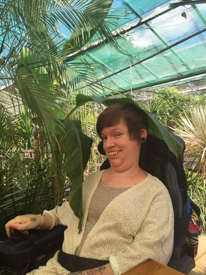 Gemma is in her powerchair and smiling, she has a large leaf hanging above her head. Gemma is at a tropical outdoor garden centre.