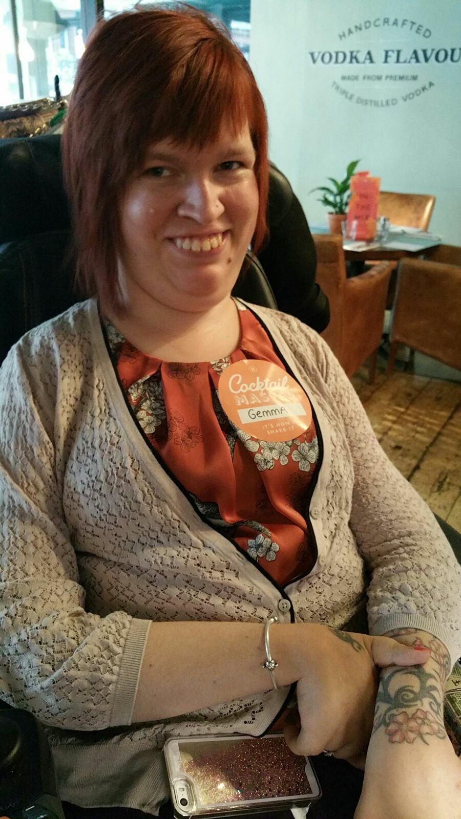 Gemma is sitting in her powerchair, smiling at the camera. She is at a cocktail making class. Gemma is also wearing a badge with her name on.
