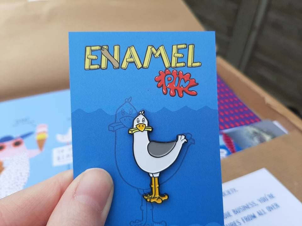 Ami is holding a small card with an enamel pin badge attached. The pin badge is a cartoon design of a seagull with a chip in between its beak.