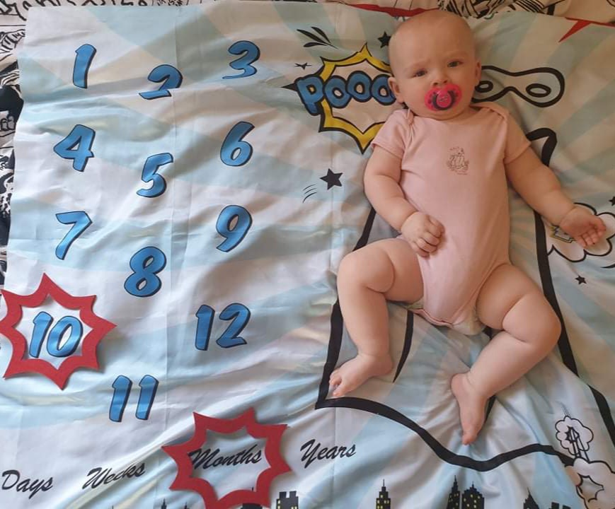 Daisy laying on her superhero-themed milestone blanket, with a red caption shape around the number 10 and another caption shape around the word months