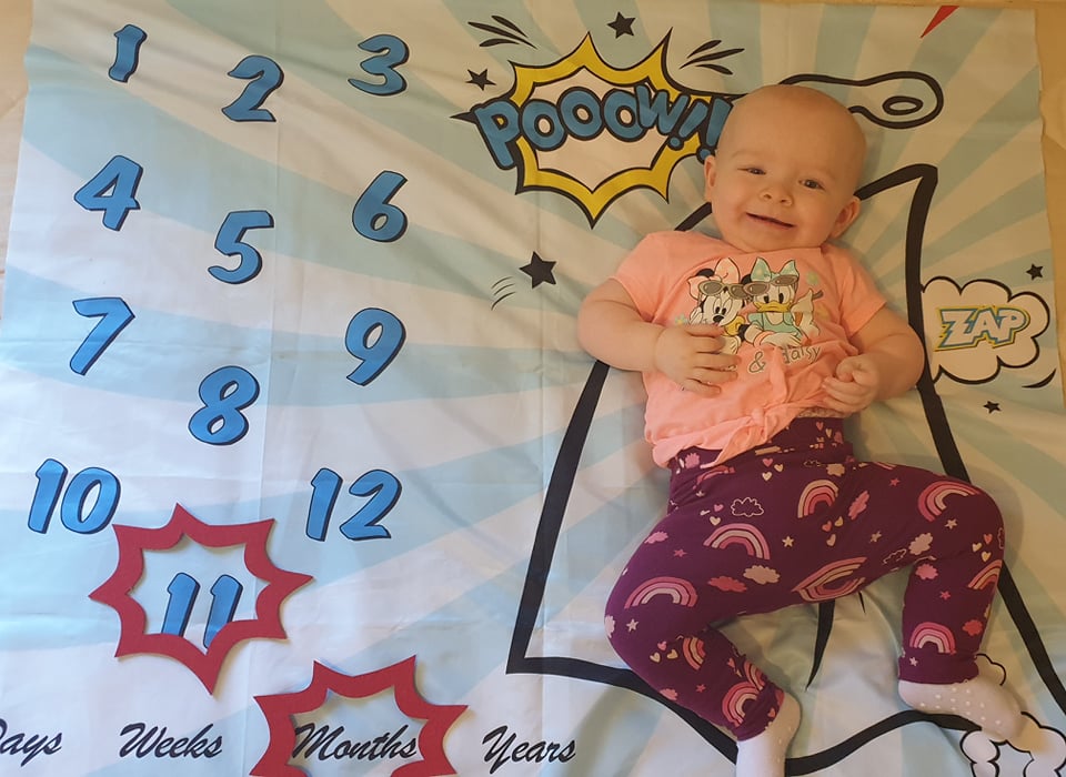 Daisy smiling, laying on her superhero-themed milestone blanket with a red caption shape around the number 11, and a red caption shape around the word months
