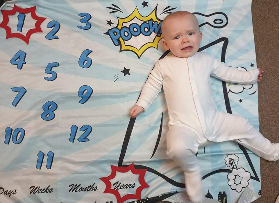 Daisy is laying on her superhero themed milestone blanket, with a red caption shape around the number 1 and another caption shape around the word years