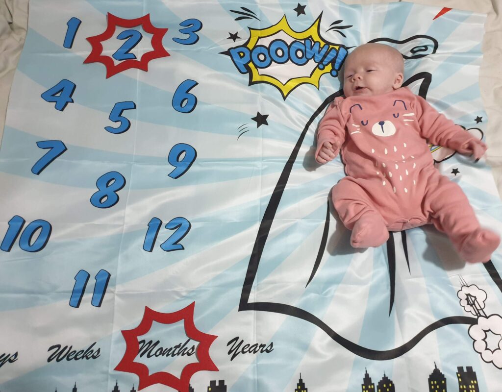 Daisy is laying on her superhero--themed milestone blanket, with a red caption shape around the number 2 and another shape around the word months