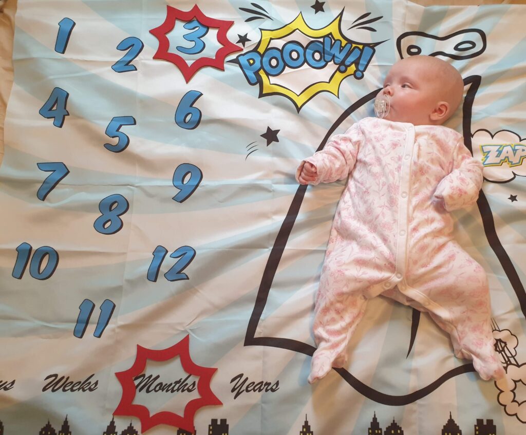 Daisy is laying on her superhero--themed milestone blanket, with a red caption shape around the number 3 and another shape around the word months