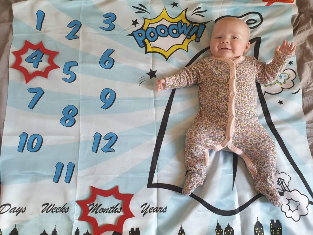 Daisy is laying on her superhero--themed milestone blanket, with a red caption shape around the number 4 and another shape around the word months