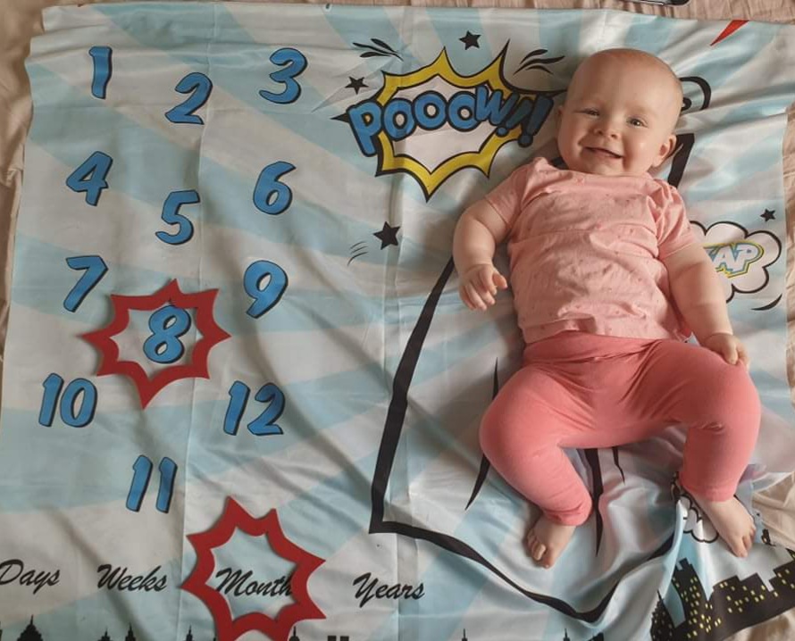 Daisy is laying on her superhero--themed milestone blanket, with a red caption shape around the number 8 and another shape around the word months