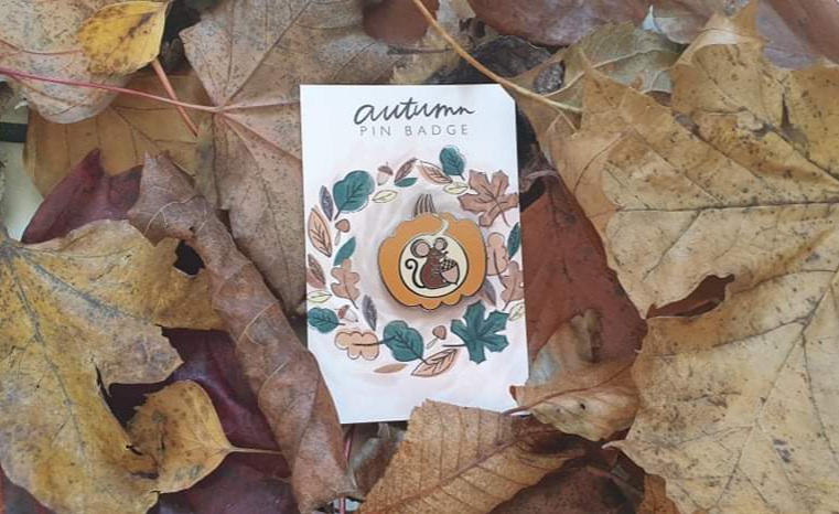 Autumn themed pin badge, an orange pumpkin with a cute little mouse inside, laid on top of leaves
