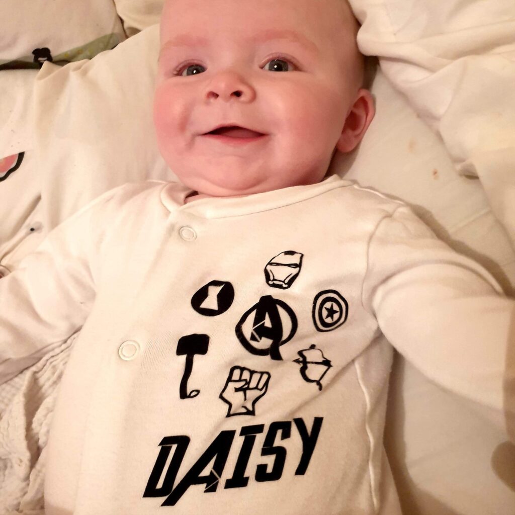 Daisy wearing a white sleepsuit, with the Avengers symbols and Daisy's name written in black