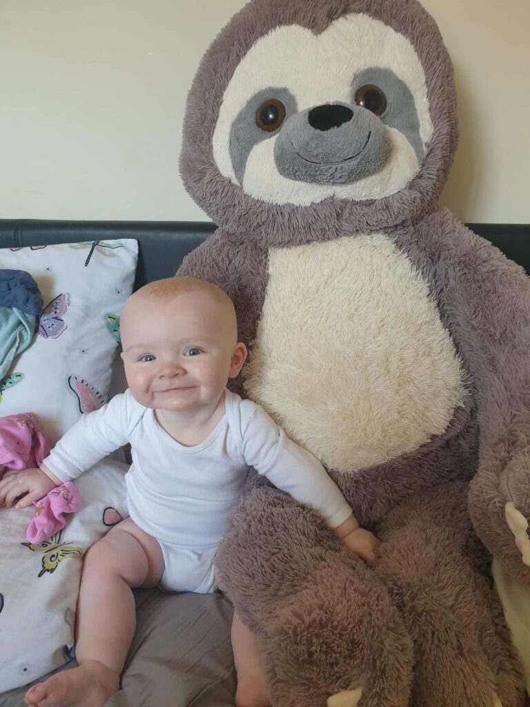 Daisy is smiling, sitting next to her very big cuddly toy, Sydney the Sloth