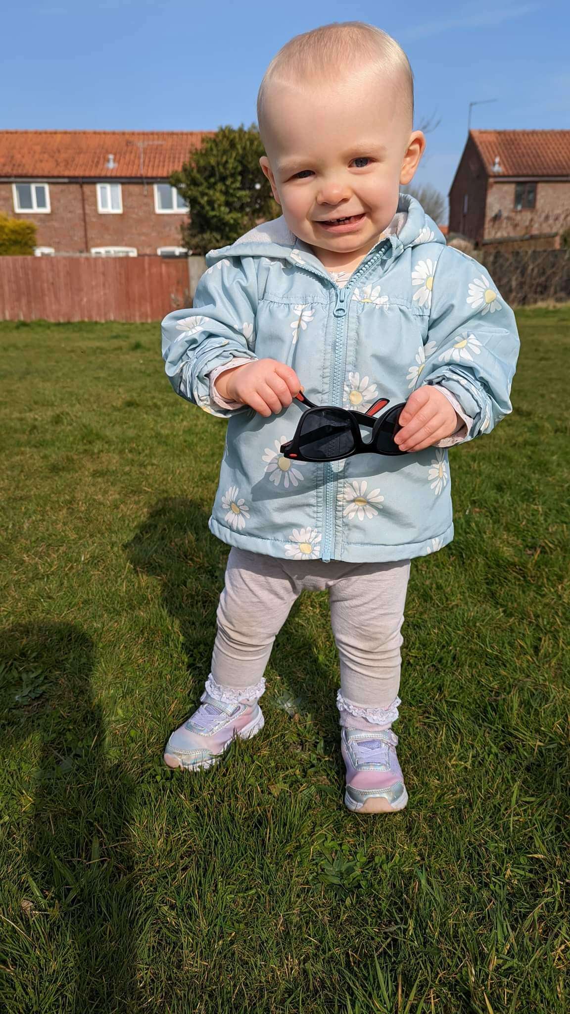 Daisy is smiling at the camera holding Ewan's sunglasses. She has very short blonde hair, blue eyes and a mischievious teethy smile. She is wearing a baby blue padded coat with daisies scattered all over the coat, with pale grey/dark white trousers, white frilly socks and pink trainers.