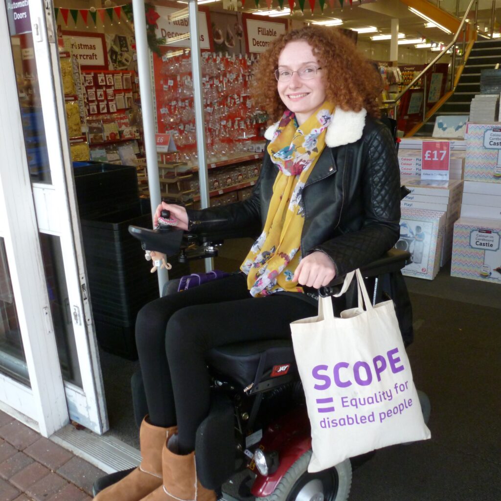 Hannah is using her powerchair, smiling whilst leaving a shop and holding a Scope bag