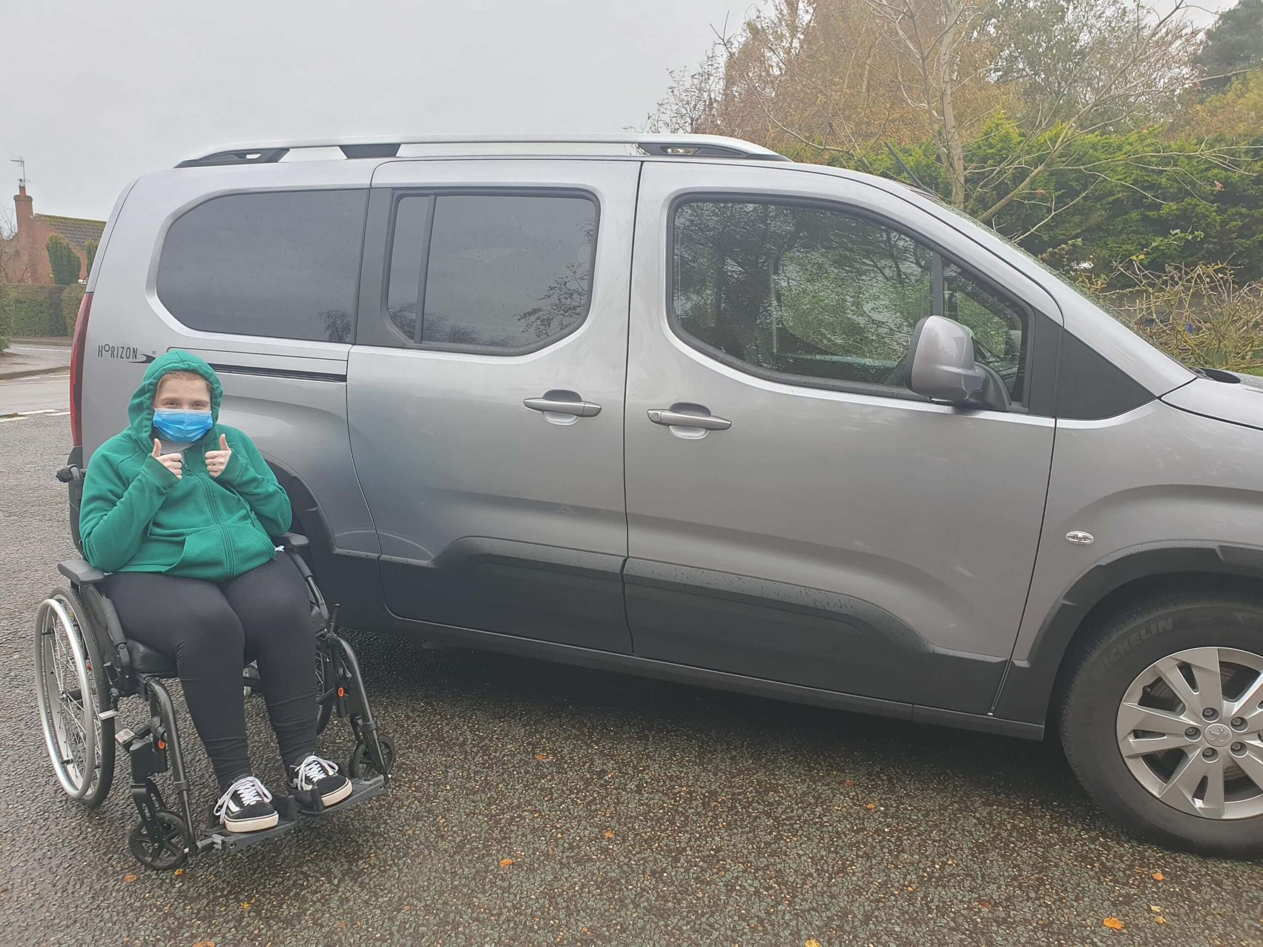 Ami is sitting in her wheelchair with both thumbs up, infront of her new WAV