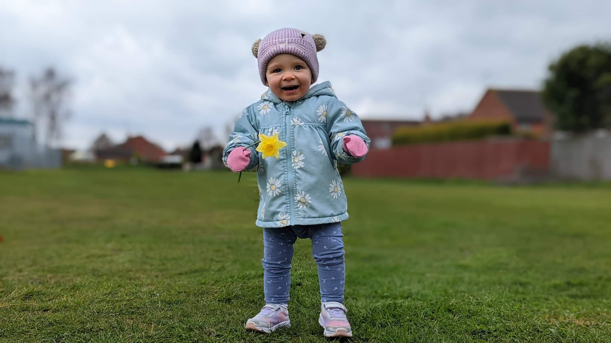 Daisy has a big smile on her face, ready to have fun at the park. She is wearing a light pink hat with a bobble on each side, a light blue padded coat with daisy pattern all over and wearing pink mittens. Also wearing light blue trousers with tiny flower patterns all over, and pink and white trainers. Daisy is holding a daffodil in her right hand.