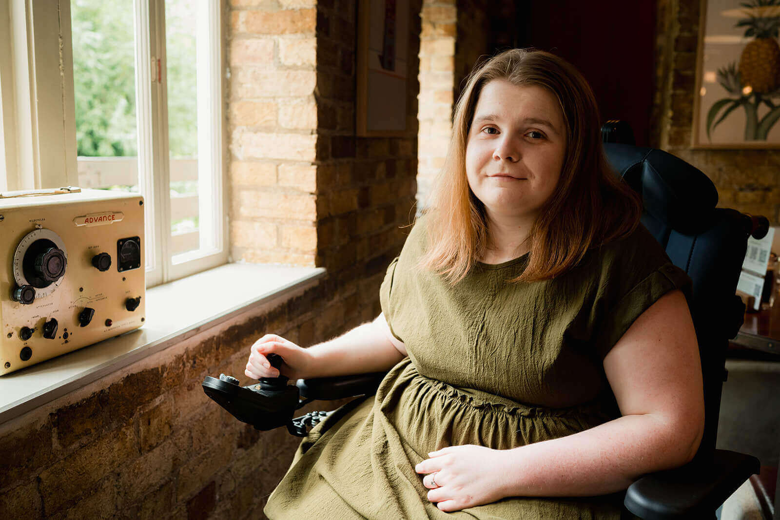 Ami is in her powerchair, smiling at the camera with the light coming through the window and shining on her. She has long brown-ginger hair and is wearing a khaki green dress.