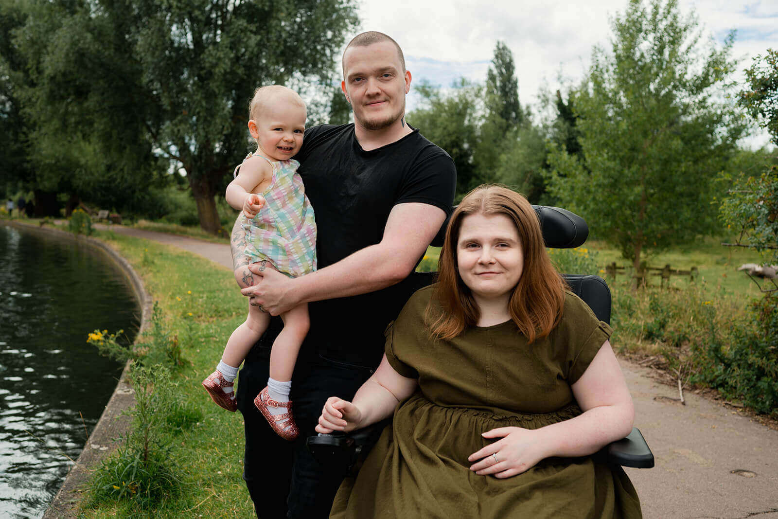 Ami is in her powerchair smiling, with Ewan standing beside her and holding Daisy. They are beside a river. Ami has long brown-ginger hair and is wearing a khaki green dress. Ewan has very short dark hair and has a light stubble. He is wearing a black t-shirt and dark blue jeans. Daisy is pointing to the camera, she has very short curly blonde hair, is wearing a lightly multi-coloured checkered dress, white frilly socks and glittery pink jelly sandals.
