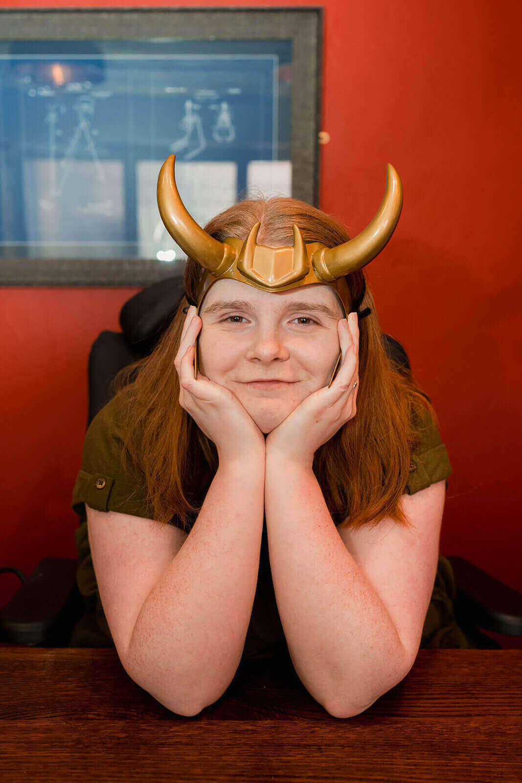 Ami is smiling, with her head rested upon the palms of her hands as her fingers cup her face. She is leaning on the table infront of her. Ami has long brown-ginger hair and is wearing a khaki green dress. She's wearing a replica of Loki's helmet.