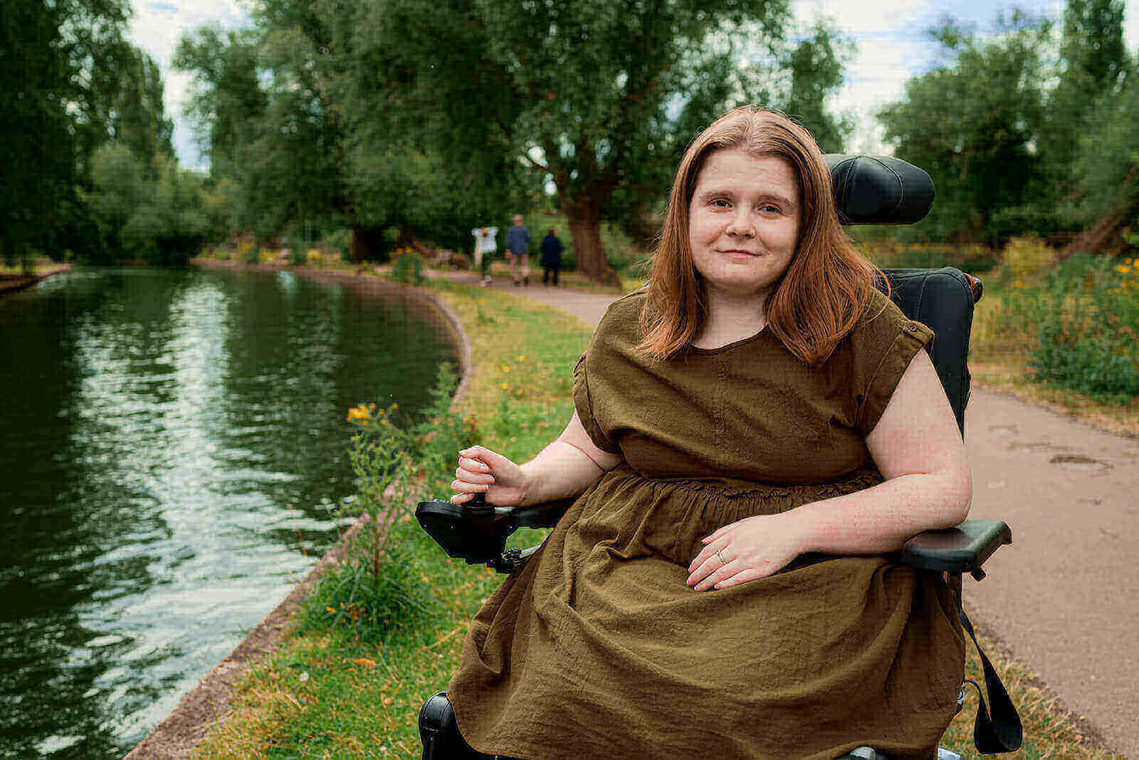 Ami is smiling, sitting in her powerchair beside a river. She has long brown-ginger hair and is wearing a khaki green dress.