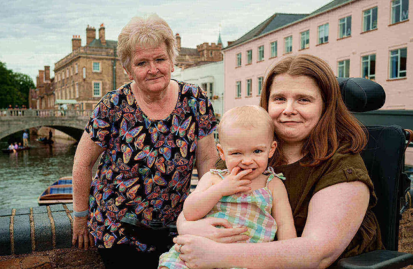 Ami is smiling, with Daisy smiling cheekily on her lap, and Ami's mum sitting beside them on a wall. Ami is in her powerchair, she has long brown-ginger hair and is wearing a khaki green dress. Daisy has very short curly blonde hair, and is wearing a lightly multi-coloured checkered dress. Ami's mum has light blonde hair and a few grey streaks (Don't tell her I said that!), is wearing a black top with multi-coloured butterflies scattered all over and black leggings. A river is in the background.